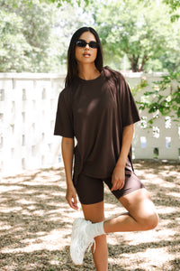 Woman in brown oversized t-shirt and biker shorts standing outdoors with a white fence and trees in the background. ::: Model is 5'10" wearing size S ///