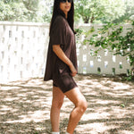 Woman in a brown outfit standing outdoors with greenery in the background