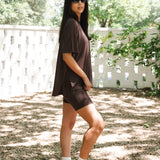 Woman in a brown outfit standing outdoors with greenery in the background