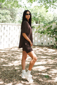 Woman in a brown outfit standing outdoors with greenery in the background