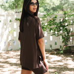 Woman wearing a brown outfit standing outdoors with trees and a white fence in the background
