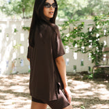 Woman wearing a brown outfit standing outdoors with trees and a white fence in the background