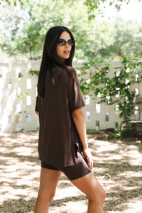Woman wearing a brown outfit standing outdoors with trees and a white fence in the background