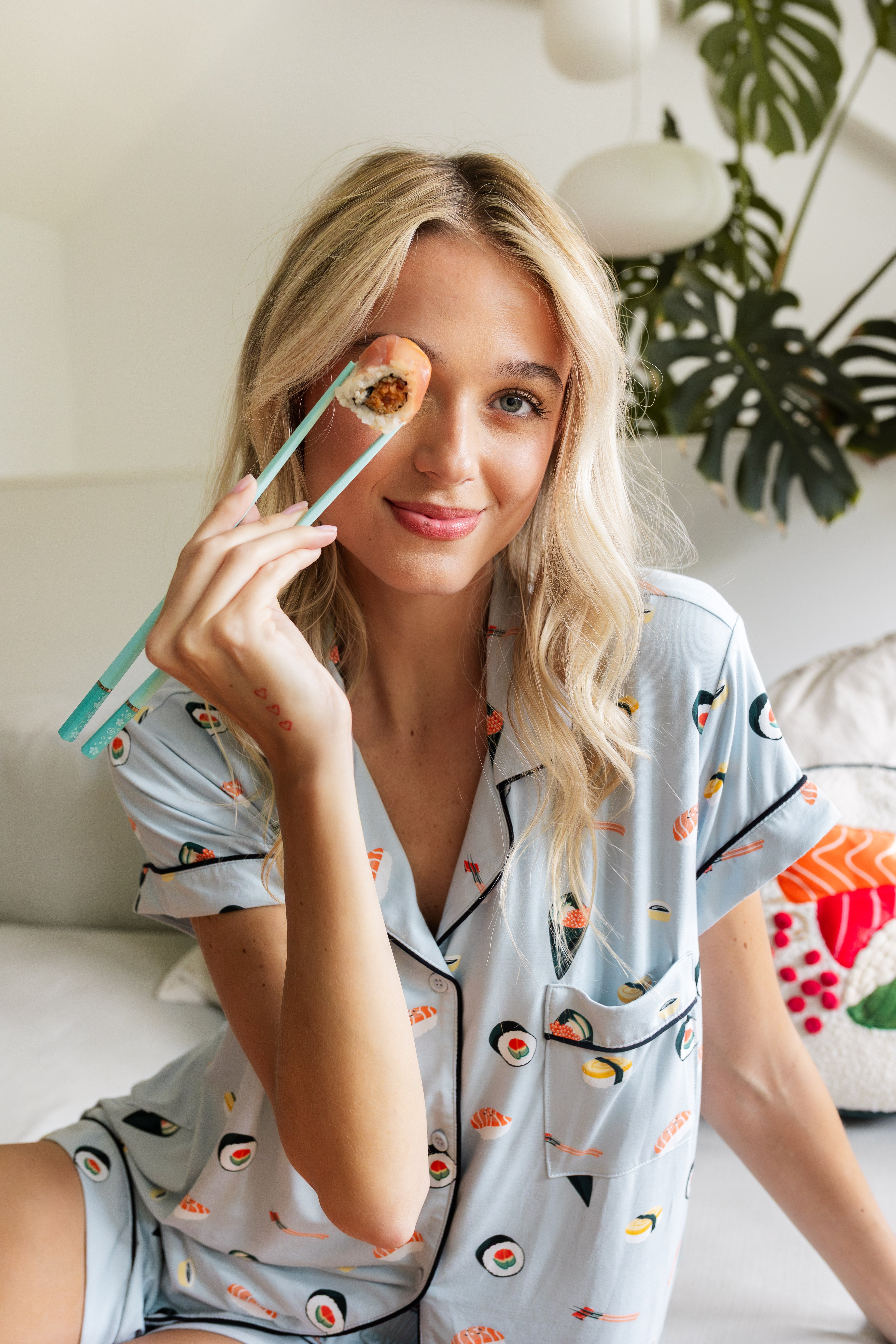 Woman holding a sushi in chopsticks wearing blue Short sleeve women's pajamas set in sushi print sitting on a white couch