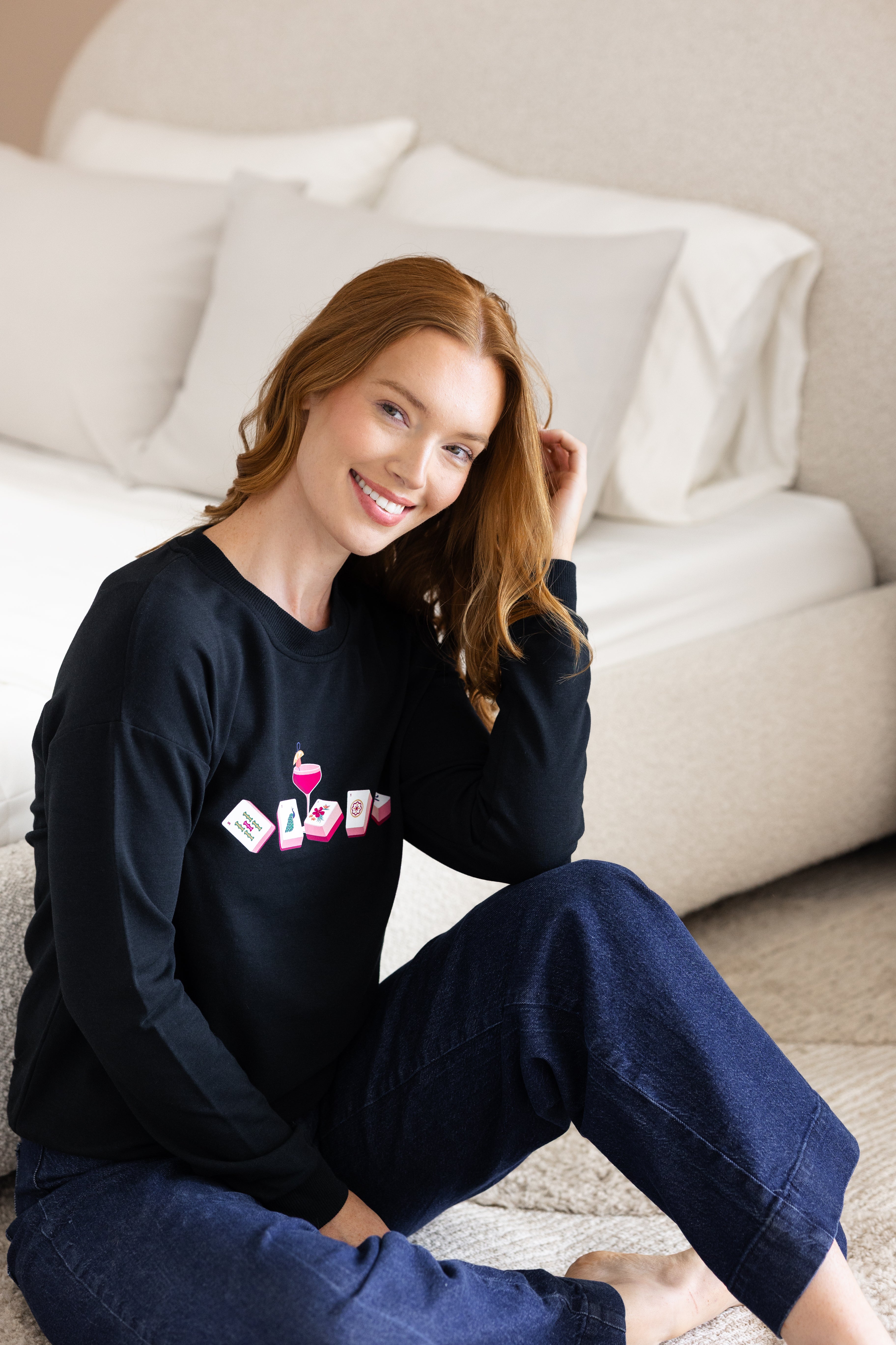 Woman wearing a black sweatshirt with colorful mahjong graphics sitting next to a bed.
