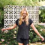 Woman in a black patterned outfit standing outdoors with greenery and a decorative wall in the background