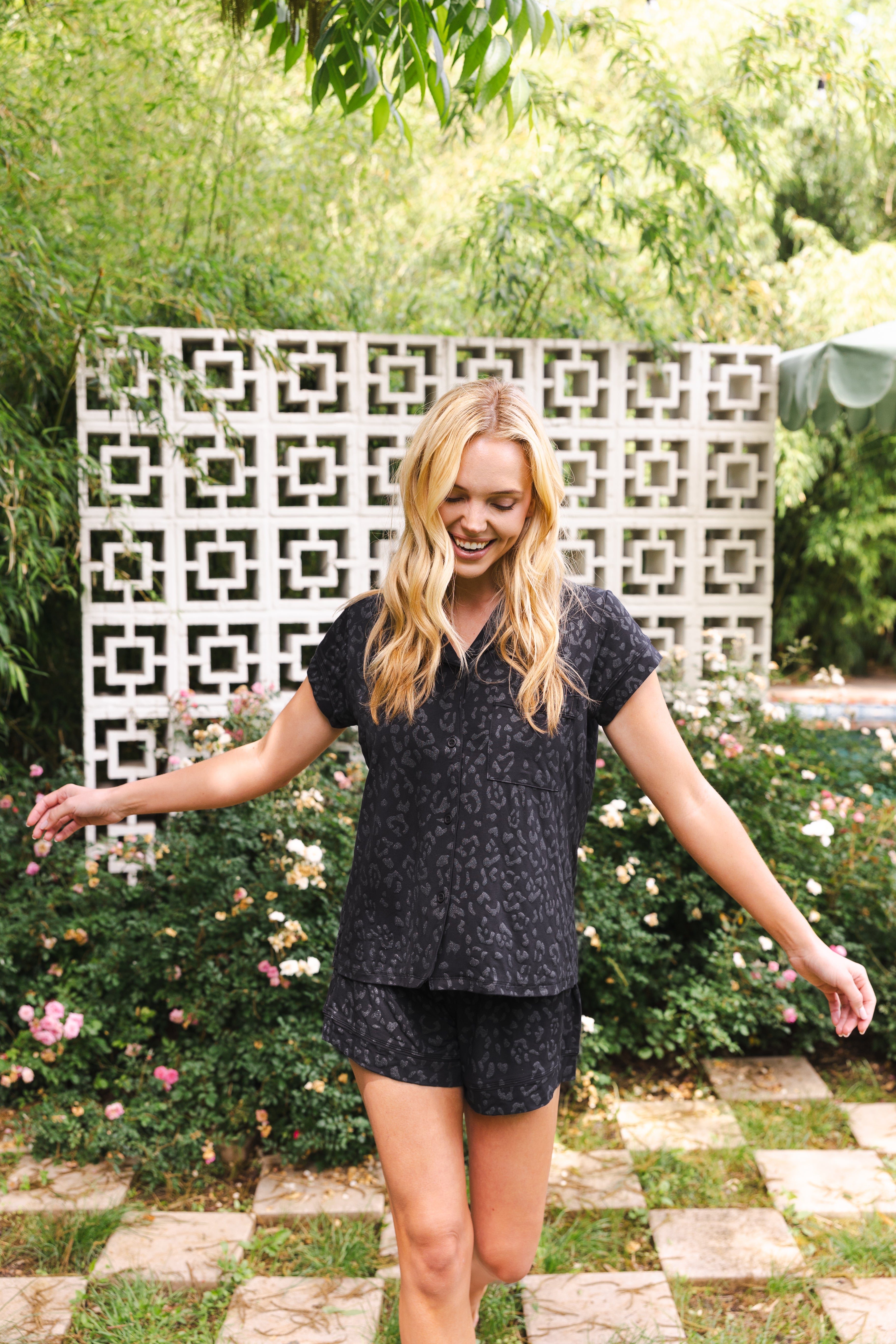 Woman in a black patterned outfit standing outdoors with greenery and a decorative wall in the background