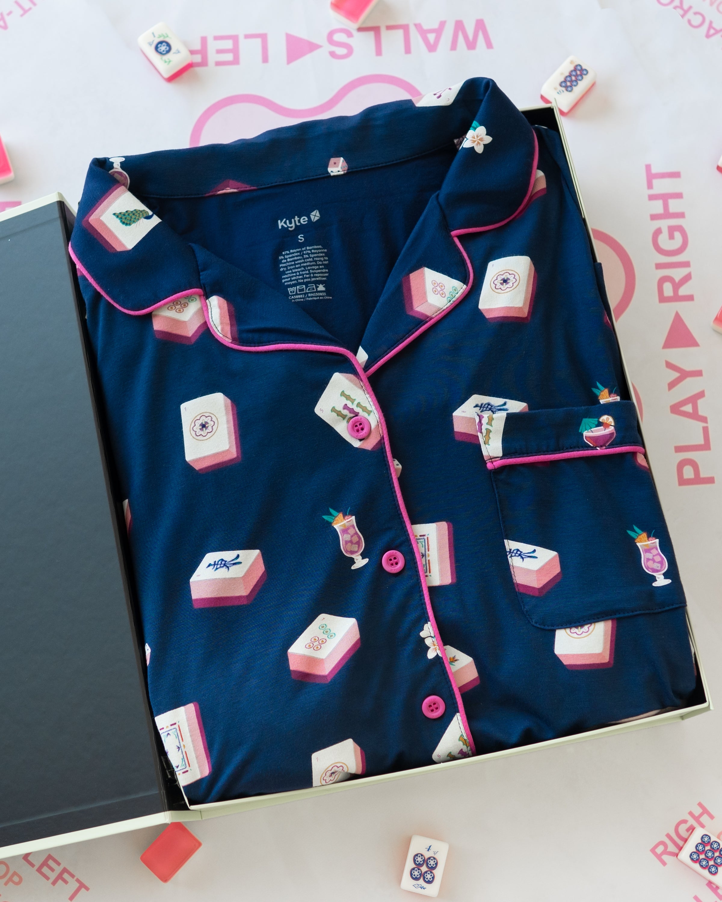 A folded navy blue pajama set with pink trim and an all-over print of pink-and-white mahjong tiles and cocktail glasses, displayed in an open box on a white background with pink text and small tile props.