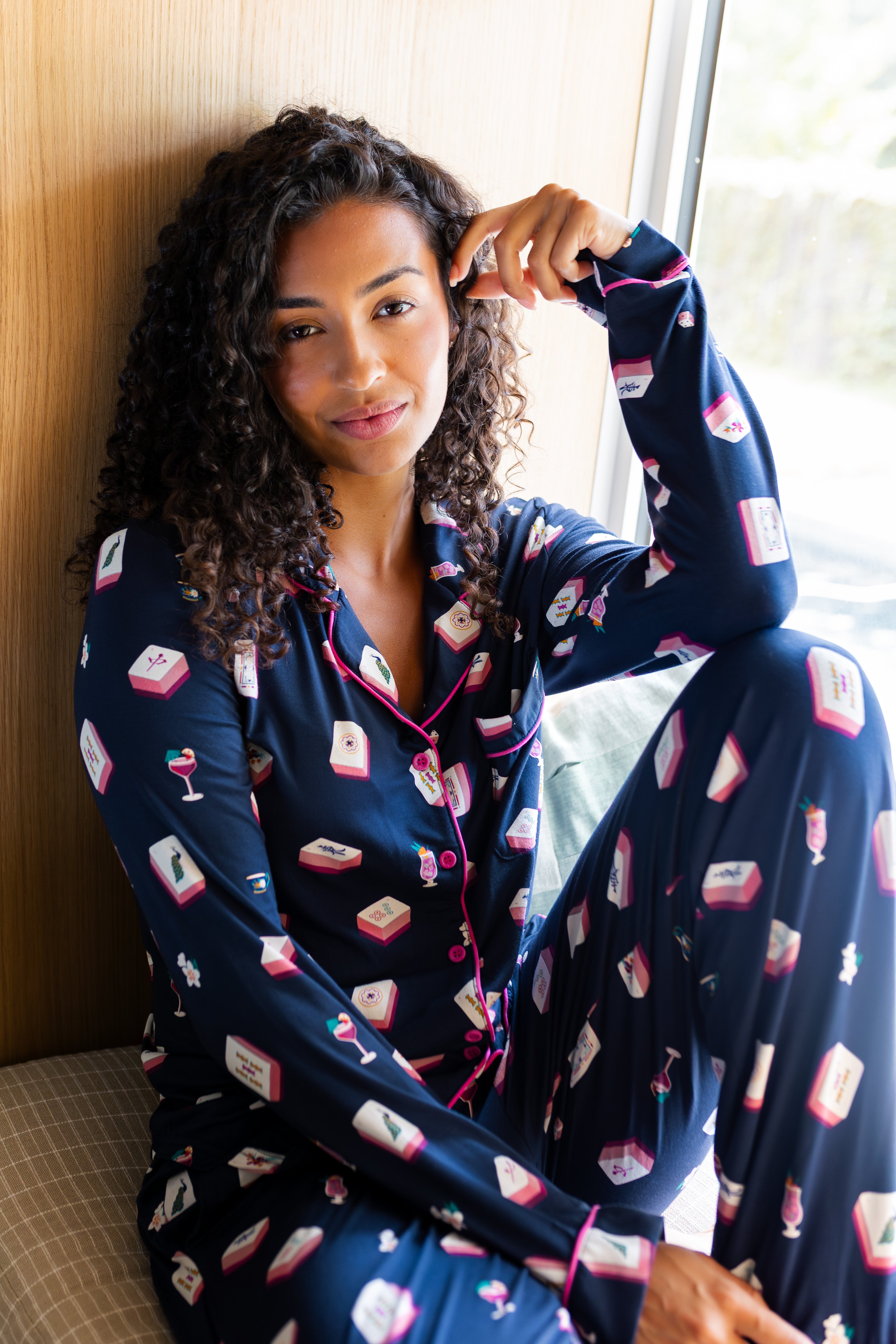 Woman wearing a navy pajama set with a mahjong pattern, sitting by a window.