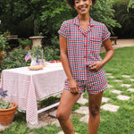 Model smiling to the side with one hand on hip while wearing the Short Sleeve Pajama Set in Patriotic Gingham with a picnic table behind her 