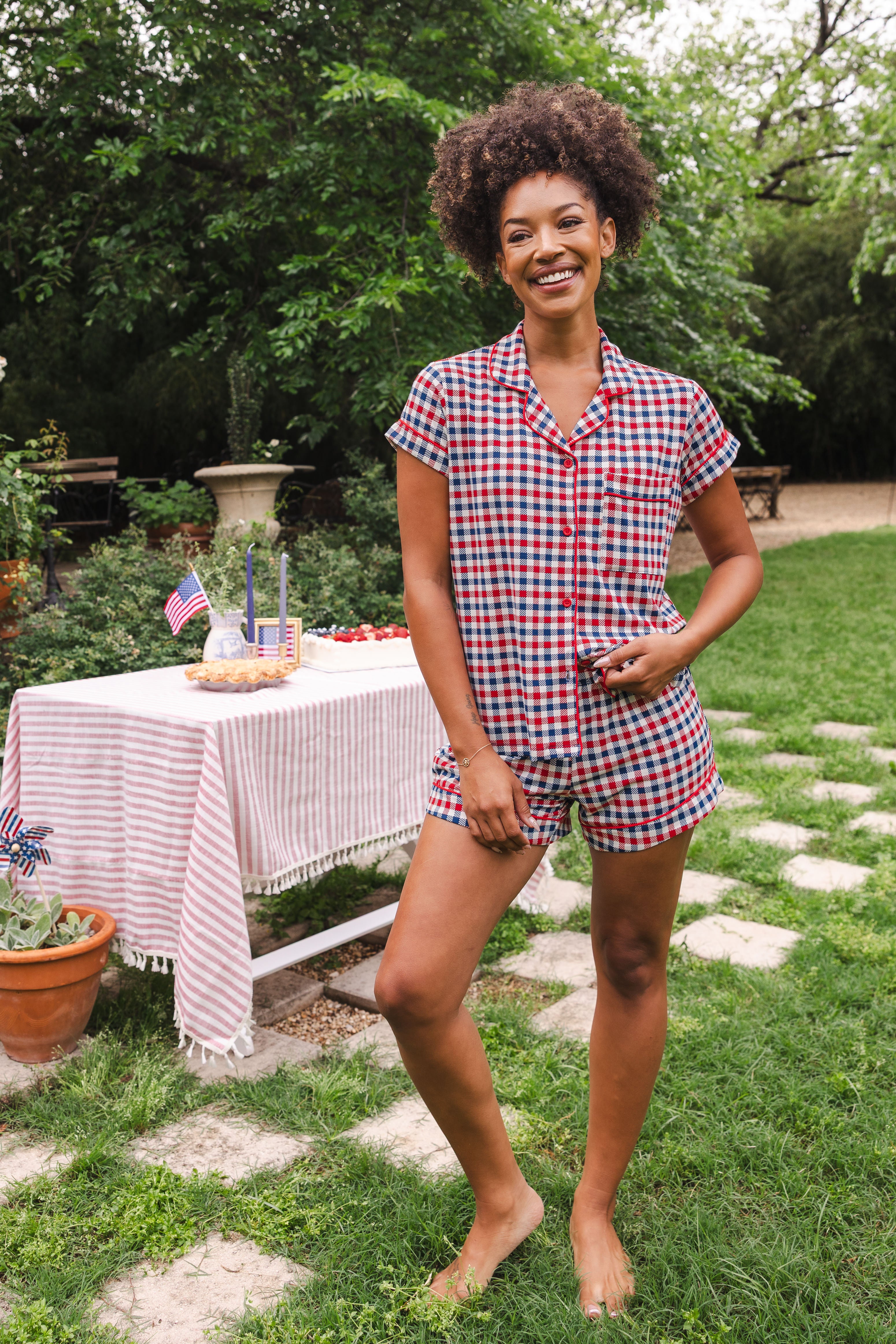 Model smiling to the side with one hand on hip while wearing the Short Sleeve Pajama Set in Patriotic Gingham with a picnic table behind her 