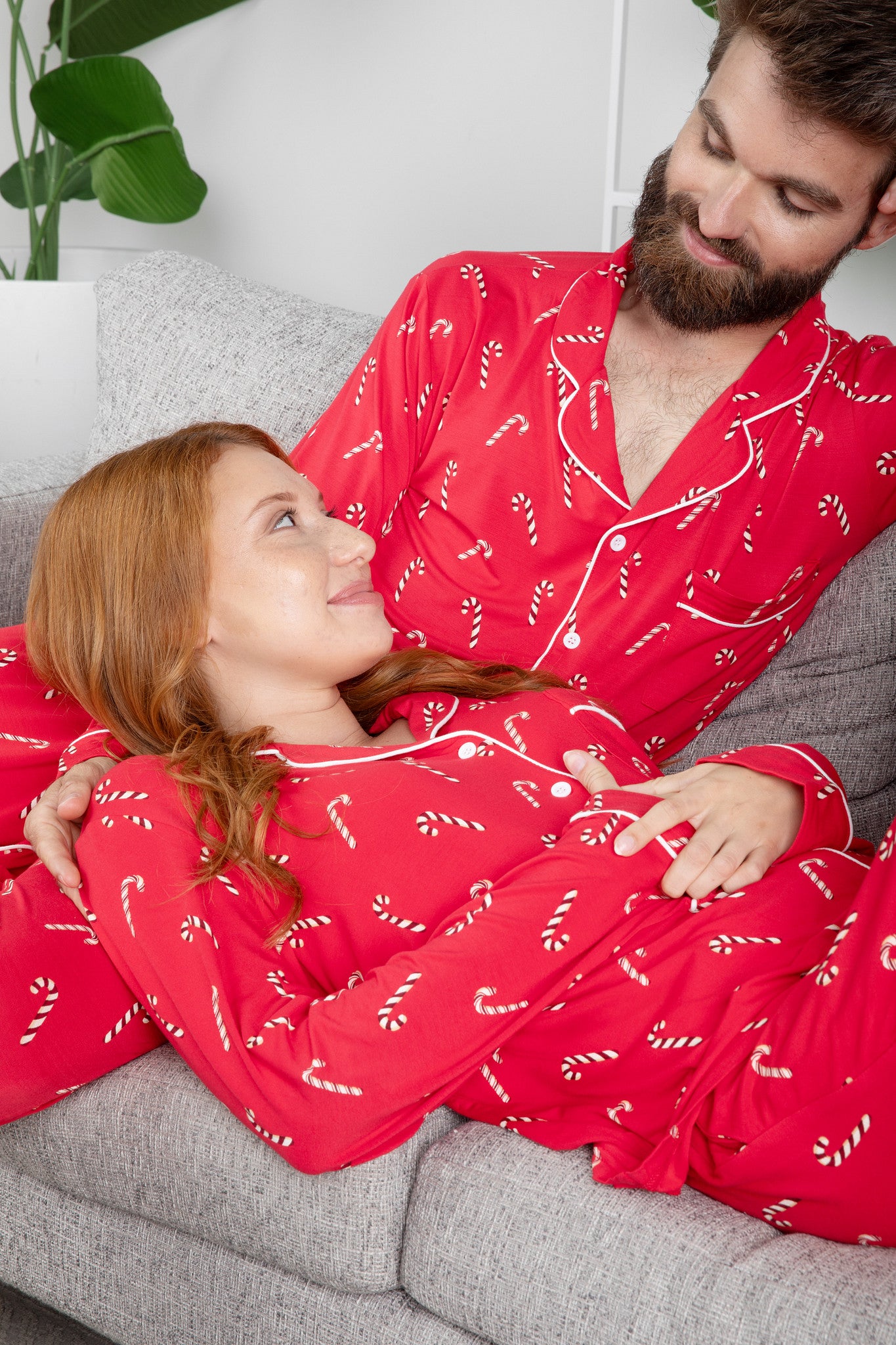 Man and woman wearing red pajamas with candy cane patterns sitting on a couch.
