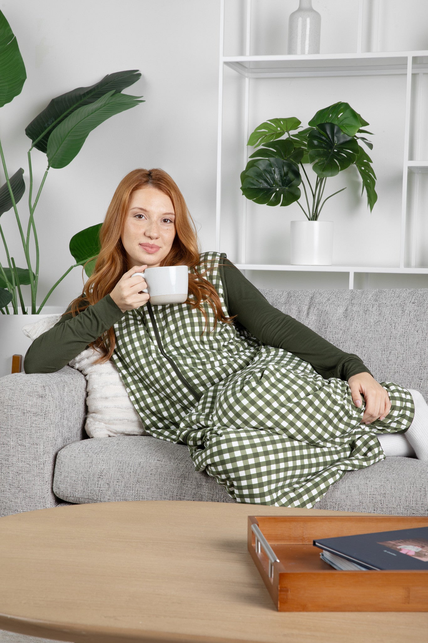 Woman in a green checkered adult sleep bag walker sitting on a couch holding a mug, with plants and a shelf in the background.