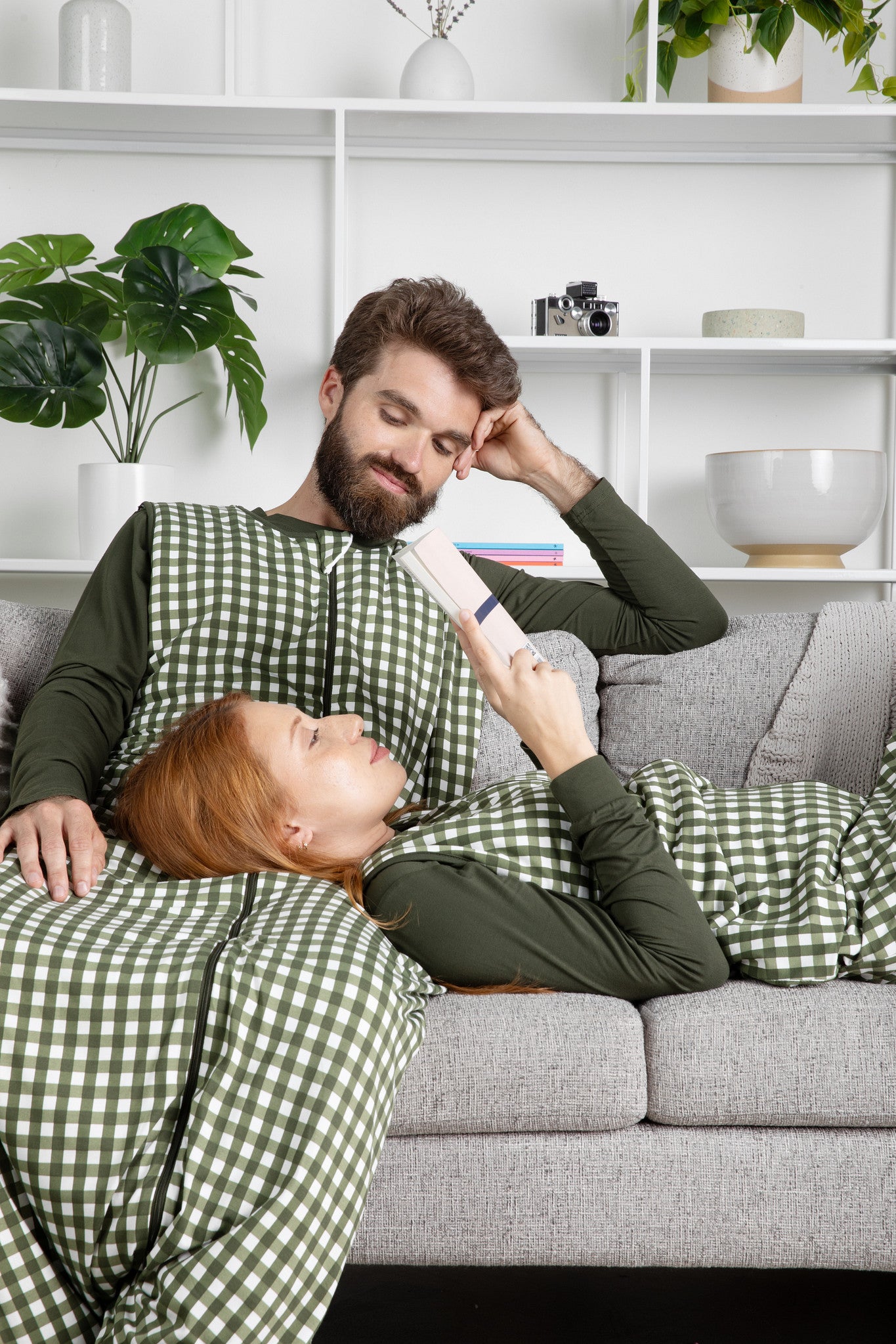 Two people in matching green checkered sleep bag walkers lying on a couch, with one person holding a book.