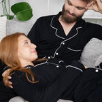 Man and woman in matching black pajamas sitting on a couch with plants in the background
