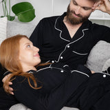 Man and woman in matching black pajamas sitting on a couch with plants in the background
