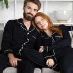 Man and woman in matching black pajamas sitting on a couch in a room with white shelves.
