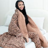 Smiling model sitting on a white bed wrapped in Kyte Quilt Blanket in Latte Leopard 2.5