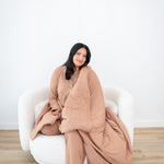 Smiling model sitting on a white sofa couch and wrapped in the Bamboo Quilt Blanket in Latte 2.5
