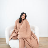 Smiling model sitting on a white sofa couch and wrapped in the Bamboo Quilt Blanket in Latte 2.5