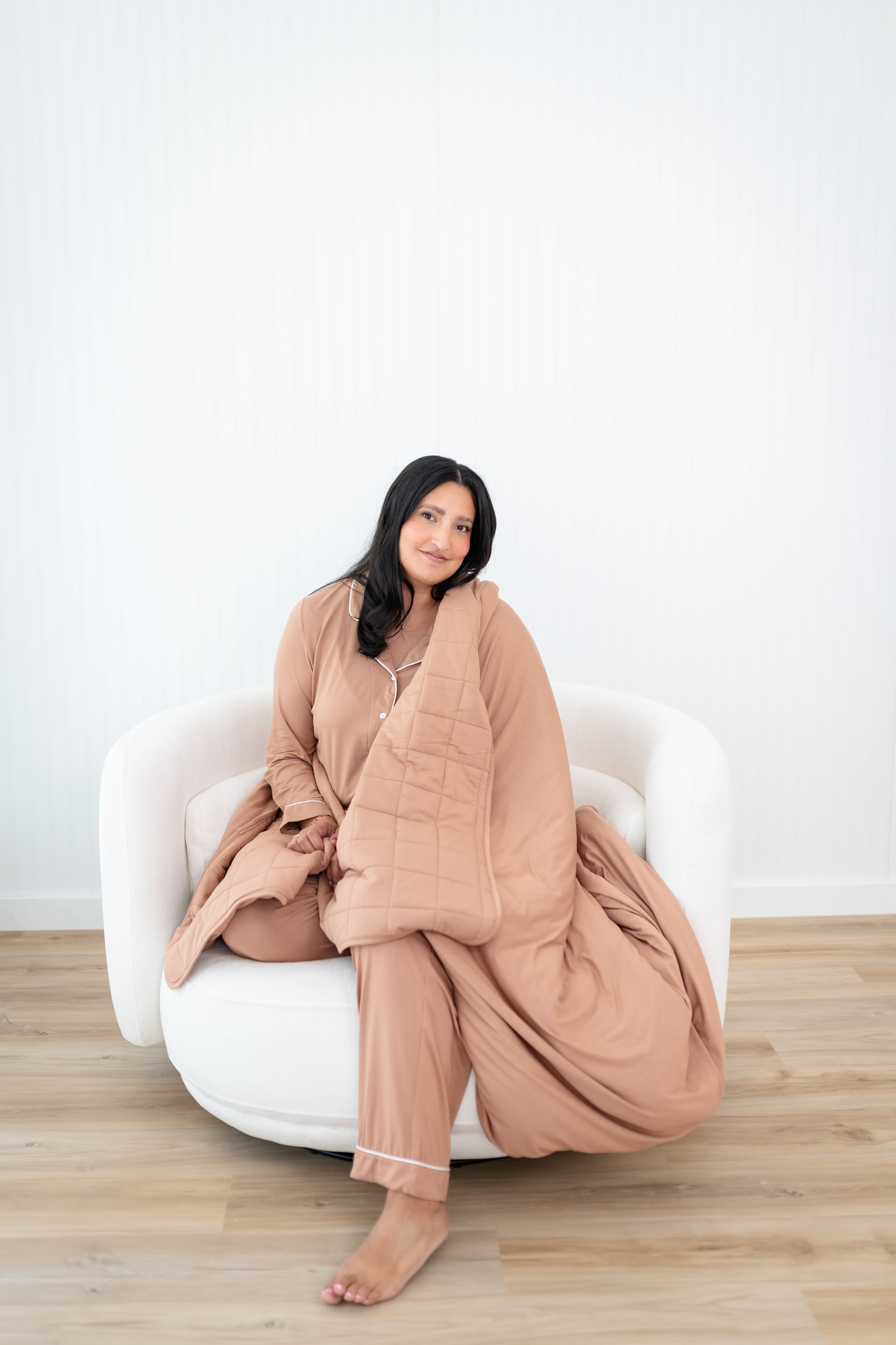 Smiling model sitting on a white sofa couch and wrapped in the Bamboo Quilt Blanket in Latte 2.5
