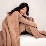 Model sitting wrapped up in the Bamboo Quilt Blanket in Latte 2.5 while wearing matching pajamas