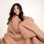 Model sitting wrapped in the Kyte Bamboo Quilt Blanket in Latte 2.5 and looking away 