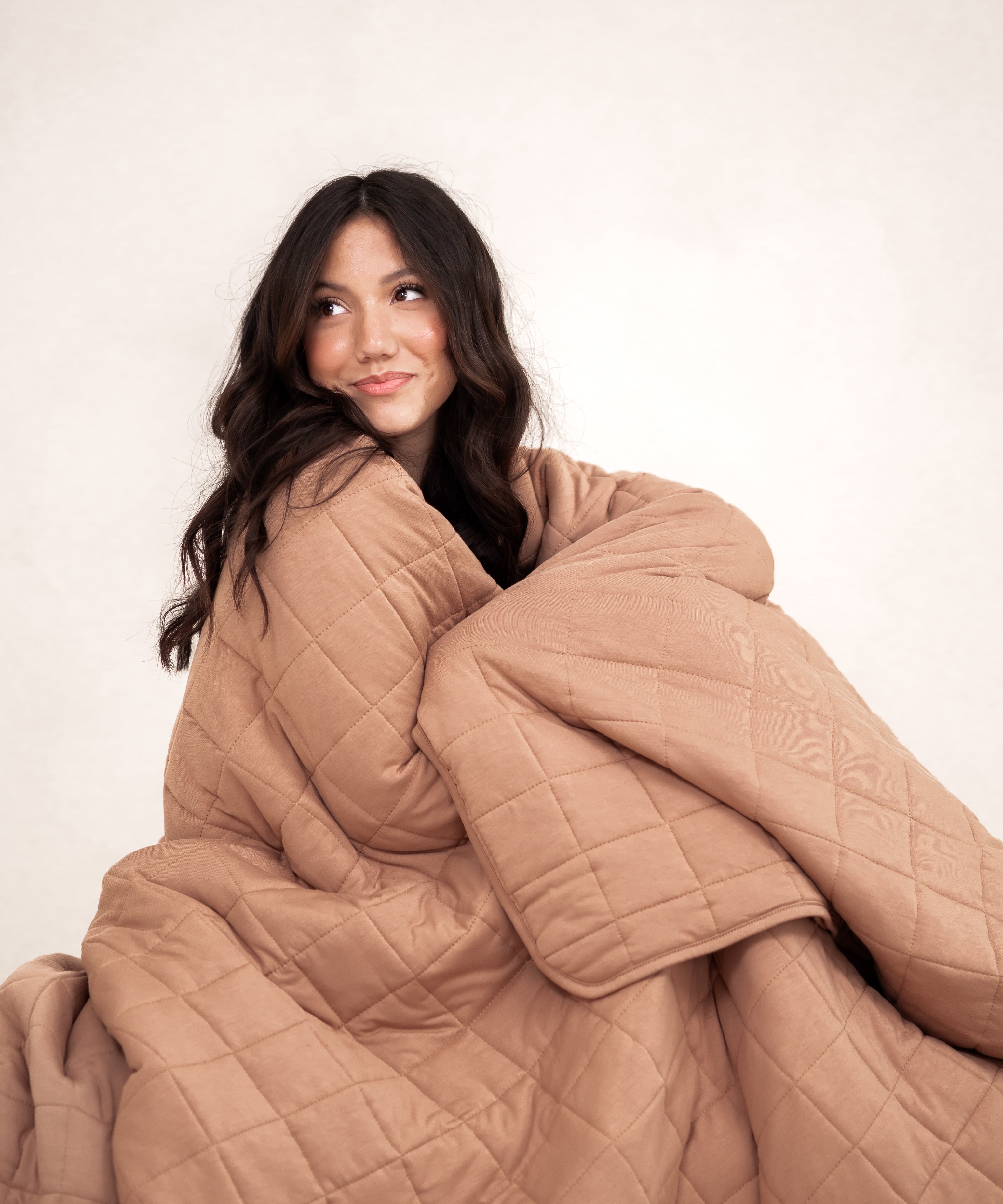 Model sitting wrapped in the Kyte Bamboo Quilt Blanket in Latte 2.5 and looking away 