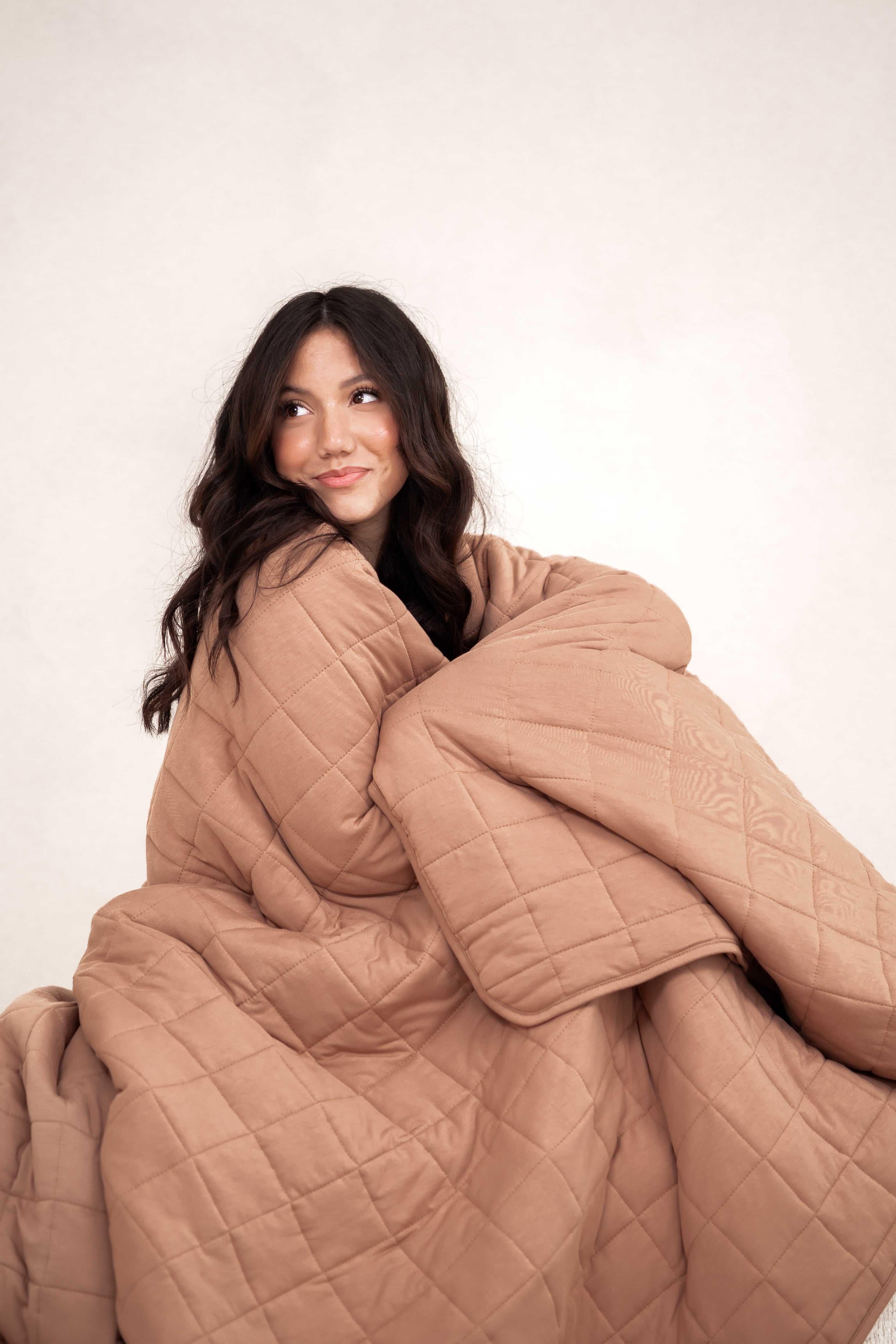 Model sitting wrapped in the Kyte Bamboo Quilt Blanket in Latte 2.5 and looking away 
