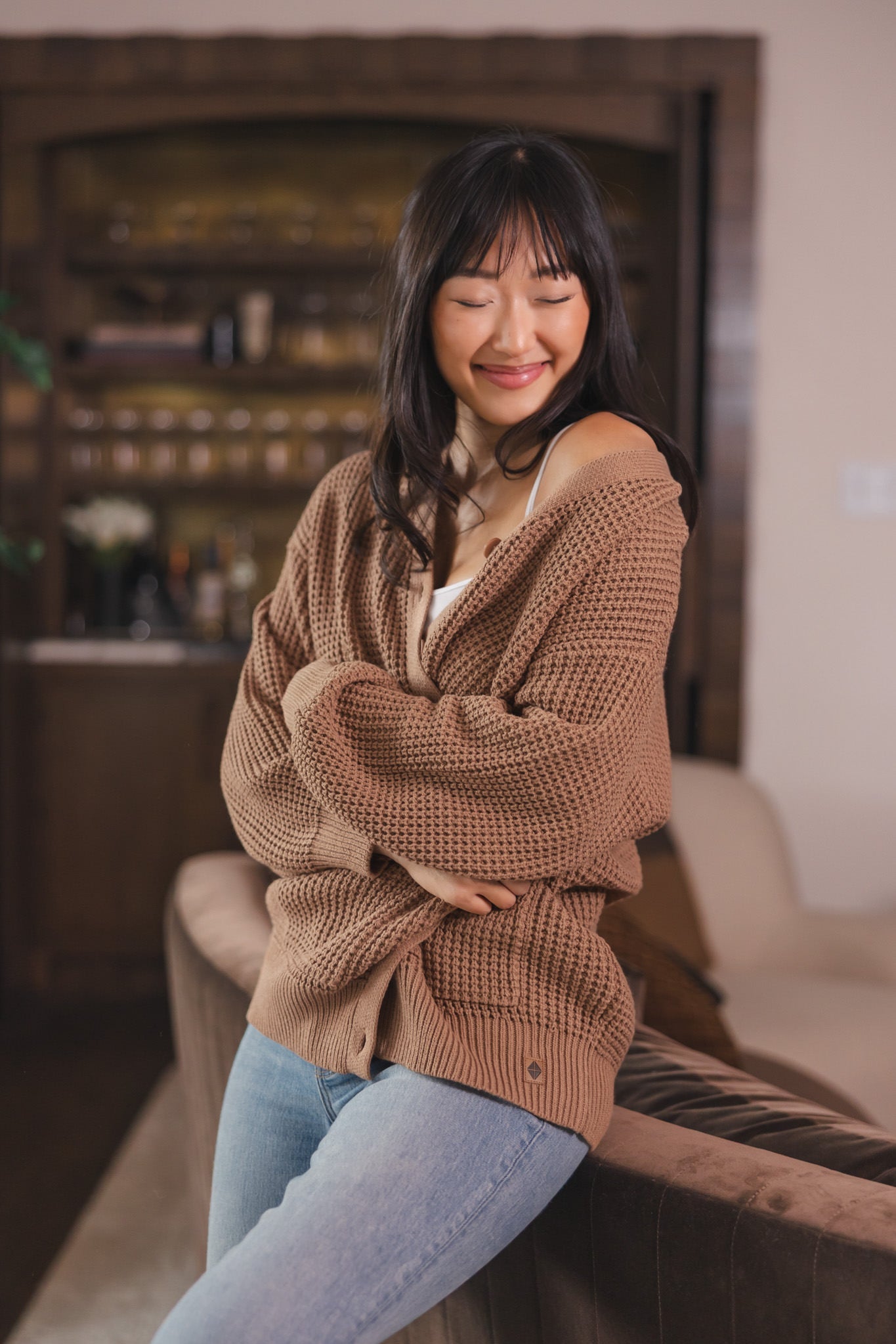Modal sitting  against a sofa in a living room wearing Chunky Knit Women's Waffle Cardigan in Latte over white camisole and blue jeans
::: Model is 5'9" wearing size S ///