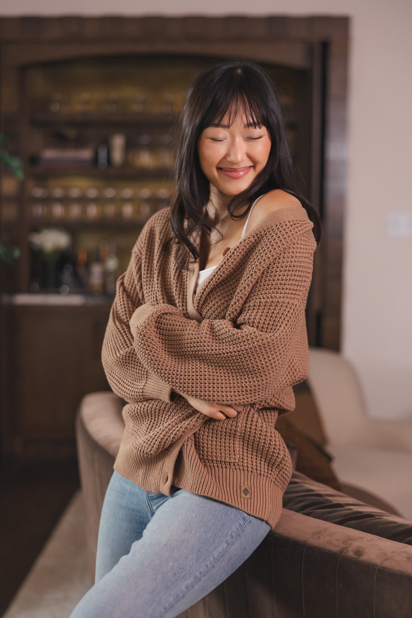 Modal sitting  against a sofa in a living room wearing Chunky Knit Women's Waffle Cardigan in Latte over white camisole and blue jeans
