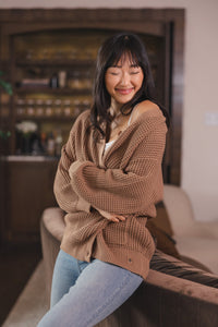 Modal sitting  against a sofa in a living room wearing Chunky Knit Women's Waffle Cardigan in Latte over white camisole and blue jeans