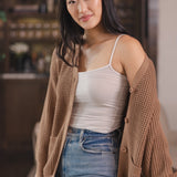 Modal standing against a sofa in a living room wearing Chunky Knit Women's Waffle Cardigan in Latte over white camisole and blue jeans