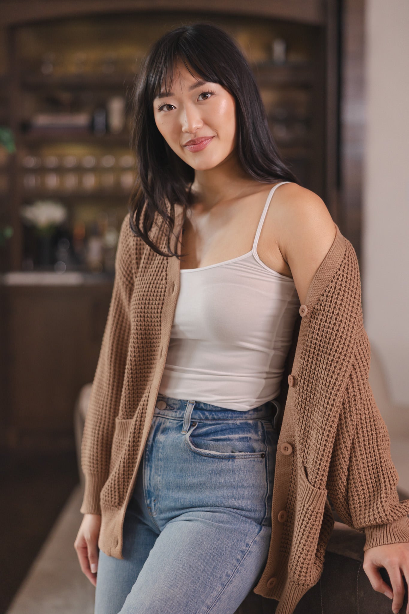 Modal standing against a sofa in a living room wearing Chunky Knit Women's Waffle Cardigan in Latte over white camisole and blue jeans