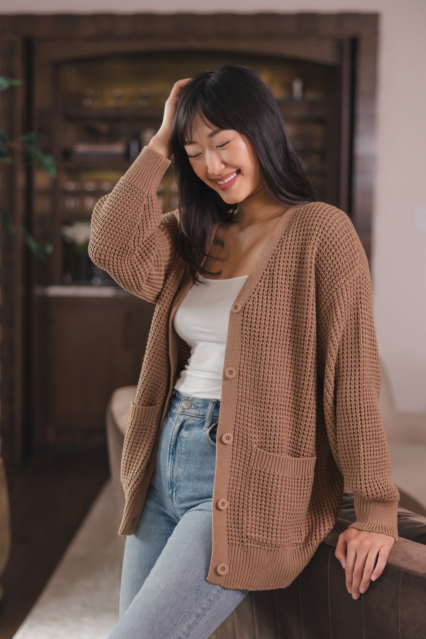 Modal smiling in a living room wearing Chunky Knit Women's Waffle Cardigan in Latte over white camisole and blue jeans