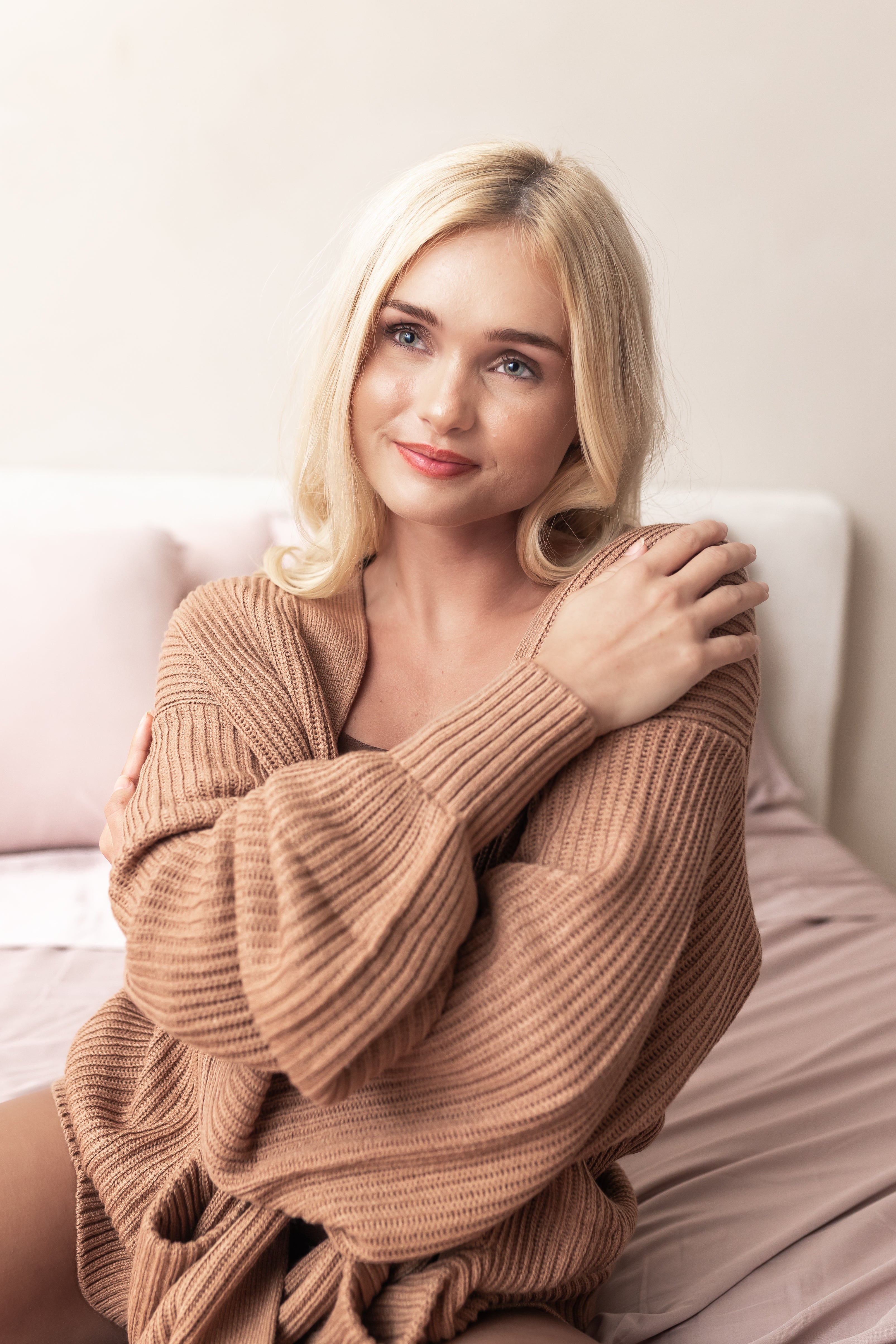 Woman wearing a warm light brown sweater sitting on a bed.