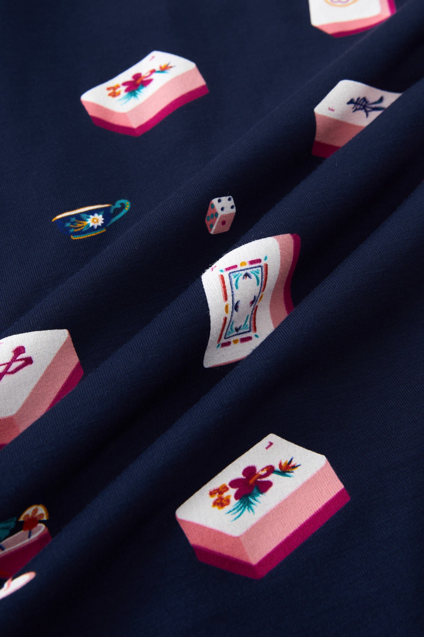 A close-up of navy blue fabric with a repeating print of Mahjong-style tiles in pink, white, and multicolor details, including floral motifs and playing card designs.