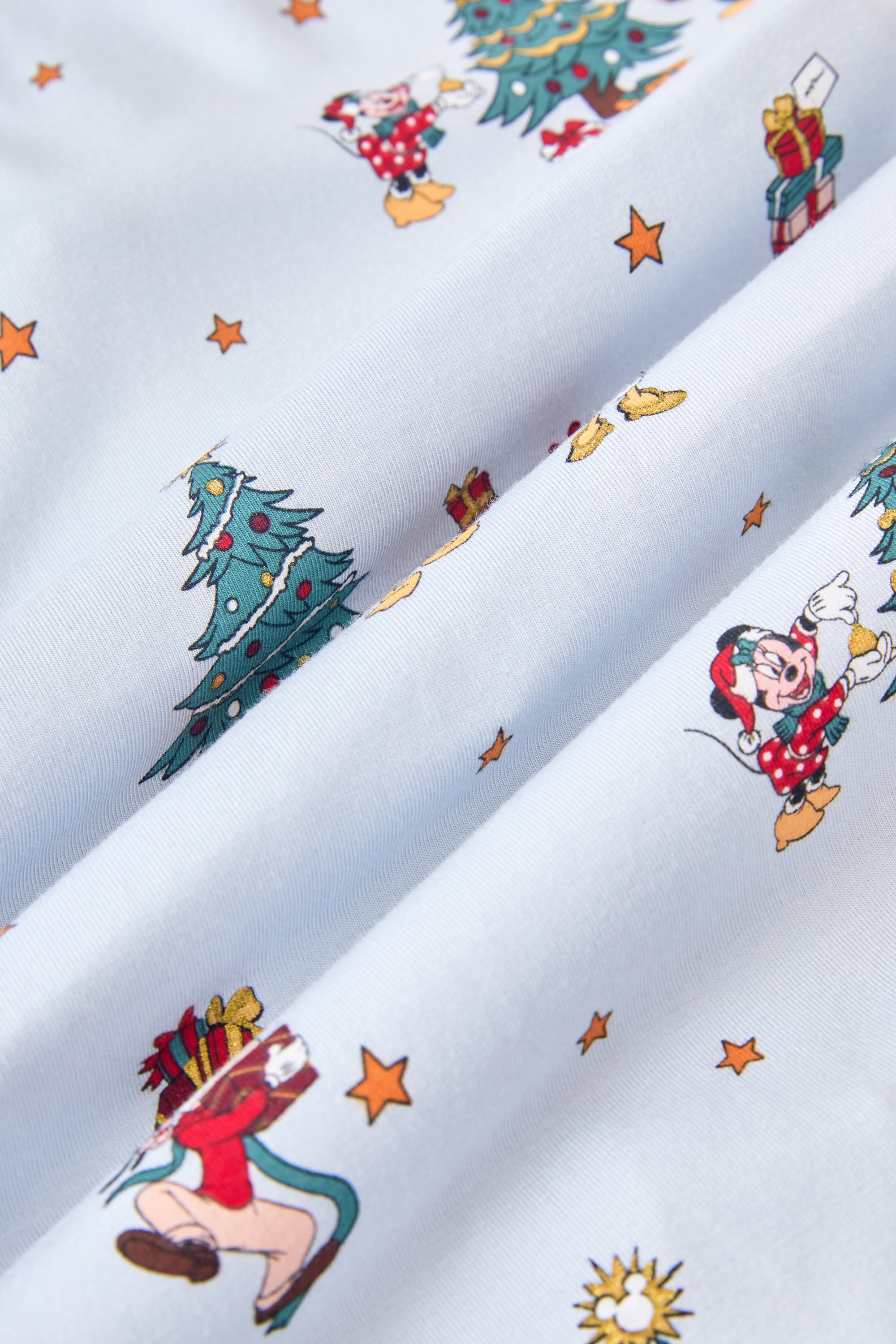 A repeating, all-over print featuring: Mickey Mouse and Minnie Mouse wearing Santa hats and decorating a Christmas tree with presents underneath; Donald Duck carrying a stack of presents away from a Christmas tree; Pluto, eyes closed and wearing a Santa hat, laying down next to a Christmas stocking filled with a bone and a candy cane; Daisy Duck holding a steaming mug; Goofy walking with a stack of presents; three wrapped presents in a stack; and gold stars scattered over a pale blue background color called