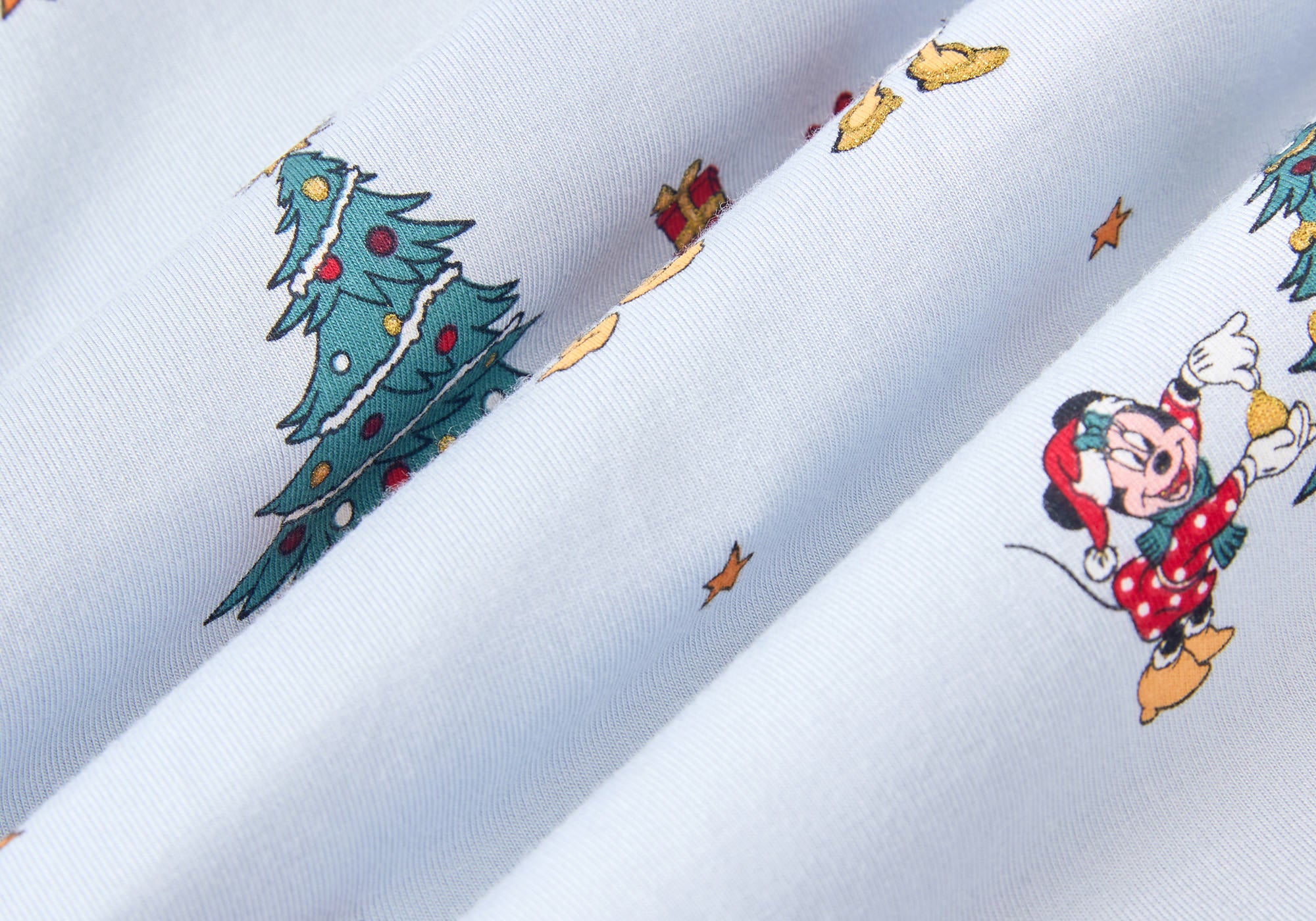 A repeating, all-over print featuring: Mickey Mouse and Minnie Mouse wearing Santa hats and decorating a Christmas tree with presents underneath; Donald Duck carrying a stack of presents away from a Christmas tree; Pluto, eyes closed and wearing a Santa hat, laying down next to a Christmas stocking filled with a bone and a candy cane; Daisy Duck holding a steaming mug; Goofy walking with a stack of presents; three wrapped presents in a stack; and gold stars scattered over a pale blue background color called