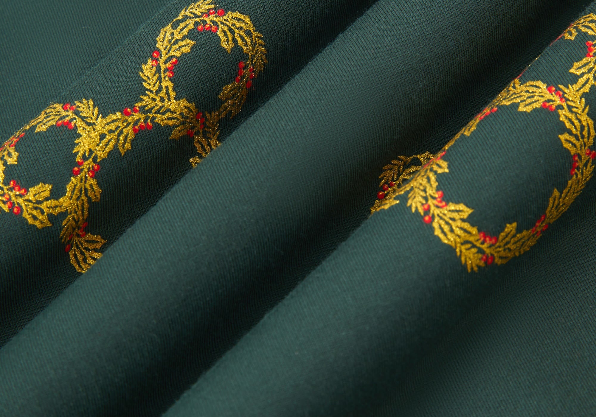 An all-over print of Mickey Mouse-shaped Christmas wreaths with gold and olive-green leaves and red holly berries, over a very dark green background color