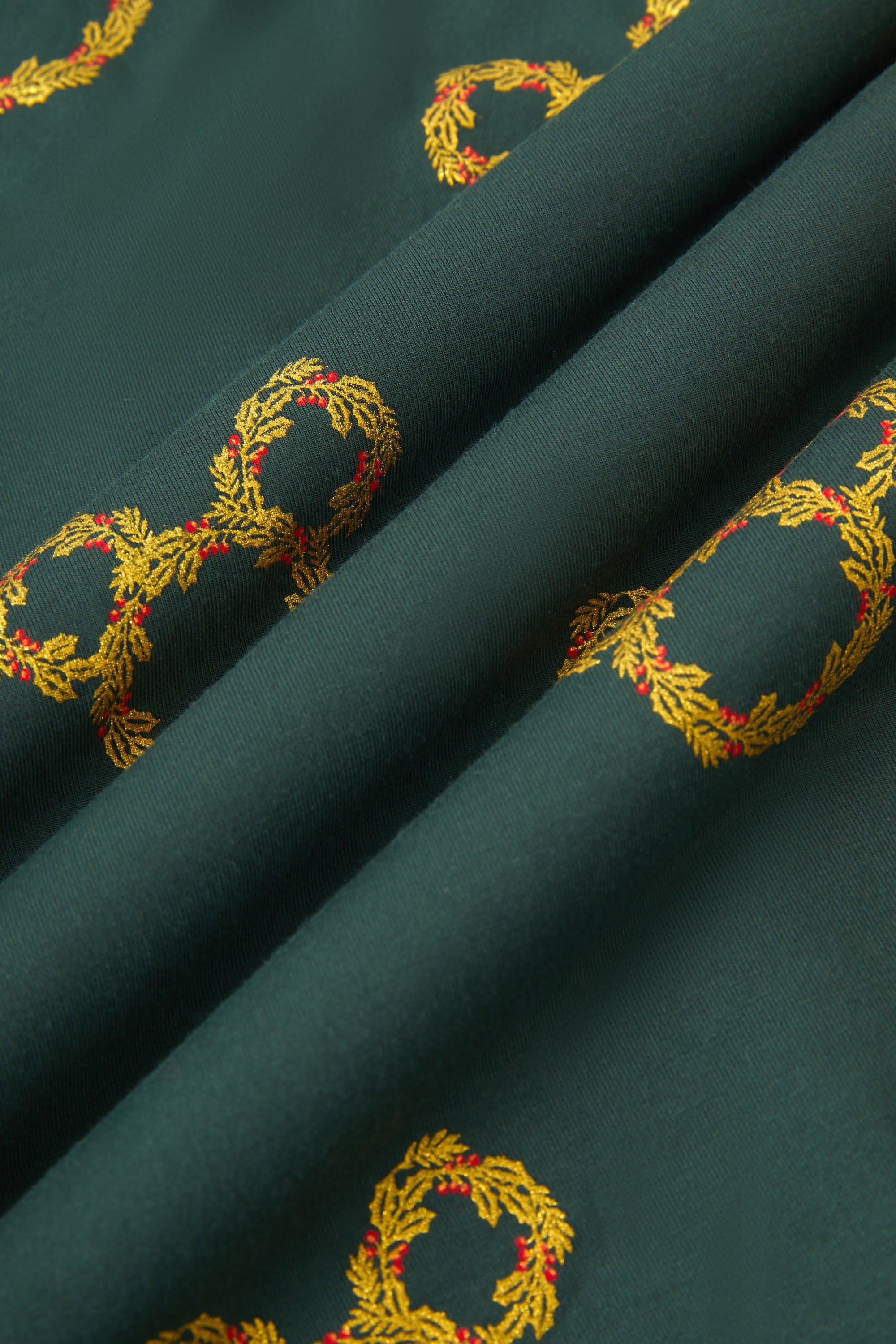 An all-over print of Mickey Mouse-shaped Christmas wreaths with gold and olive-green leaves and red holly berries, over a very dark green background color