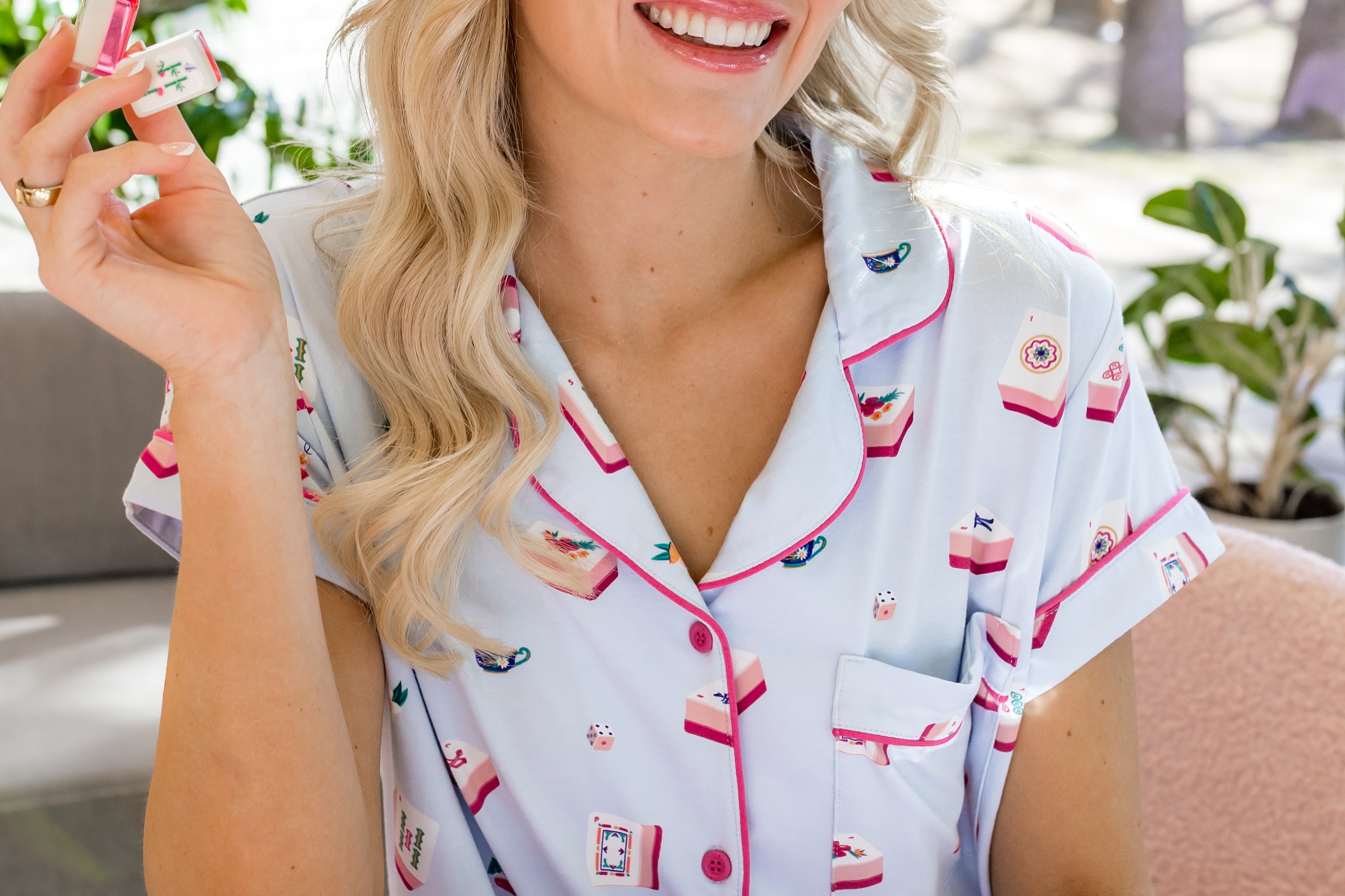 Woman in mahjong printed pajamas holding a mahjong tile, sitting at a table with pink and white items.