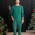 Smiling male model standing between two decorated Christmas trees wearing the Men's Lounge Pants in Merry and Bright paired with an Emerald long sleeve Henley
