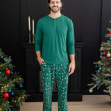 Smiling male model standing between two decorated Christmas trees wearing the Men's Lounge Pants in Merry and Bright paired with an Emerald long sleeve Henley