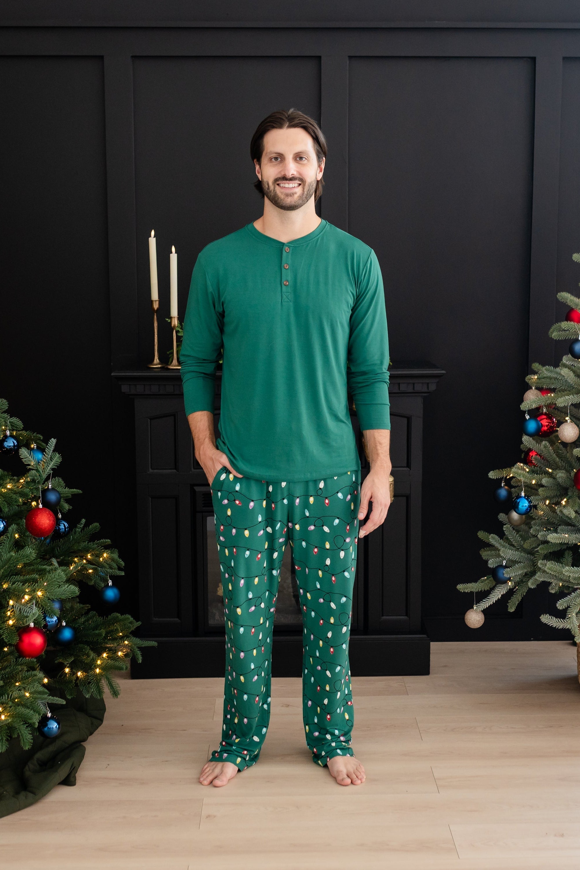 Smiling male model standing between two decorated Christmas trees wearing the Men's Lounge Pants in Merry and Bright paired with an Emerald long sleeve Henley