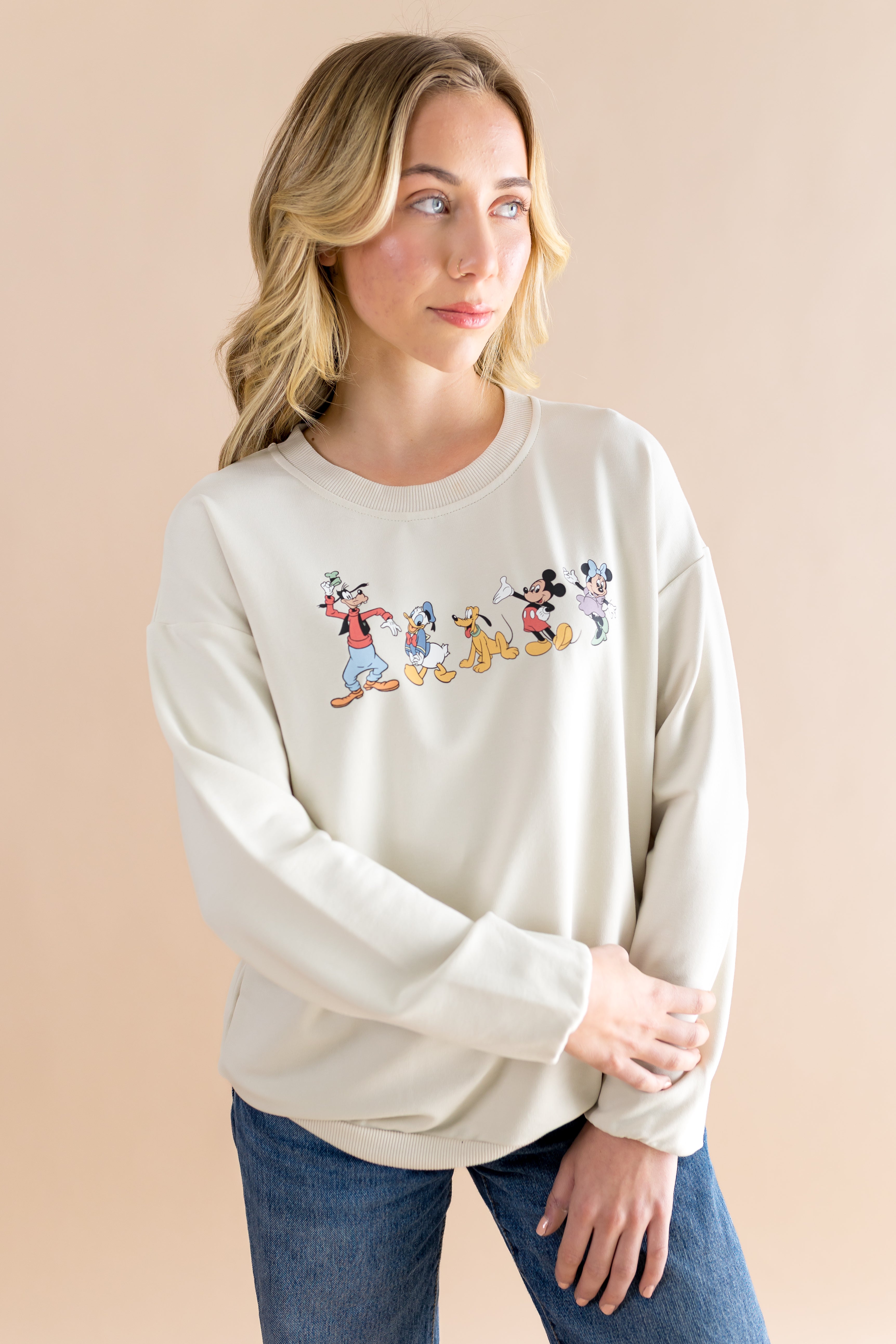 Woman standing in front of a beige back ground looking to the side while modeling the Women's Sweatshirt in Mickey and Friends on Ecru paired with dark wash jeans 
