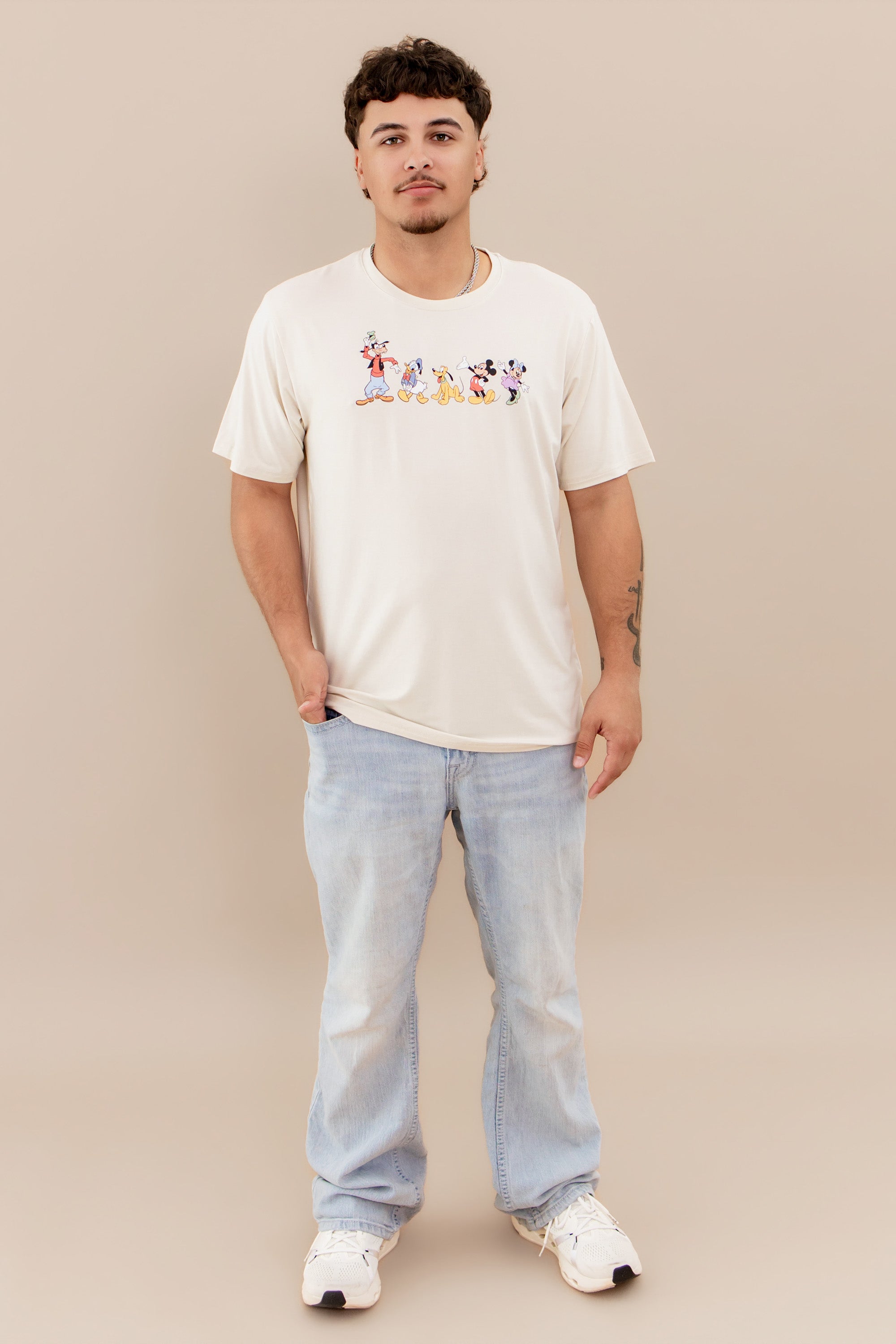 Male model wearing the Men's Crew Neck Tee in Mickey and Friends on Ecru paired with light wash jeans and white sneakers