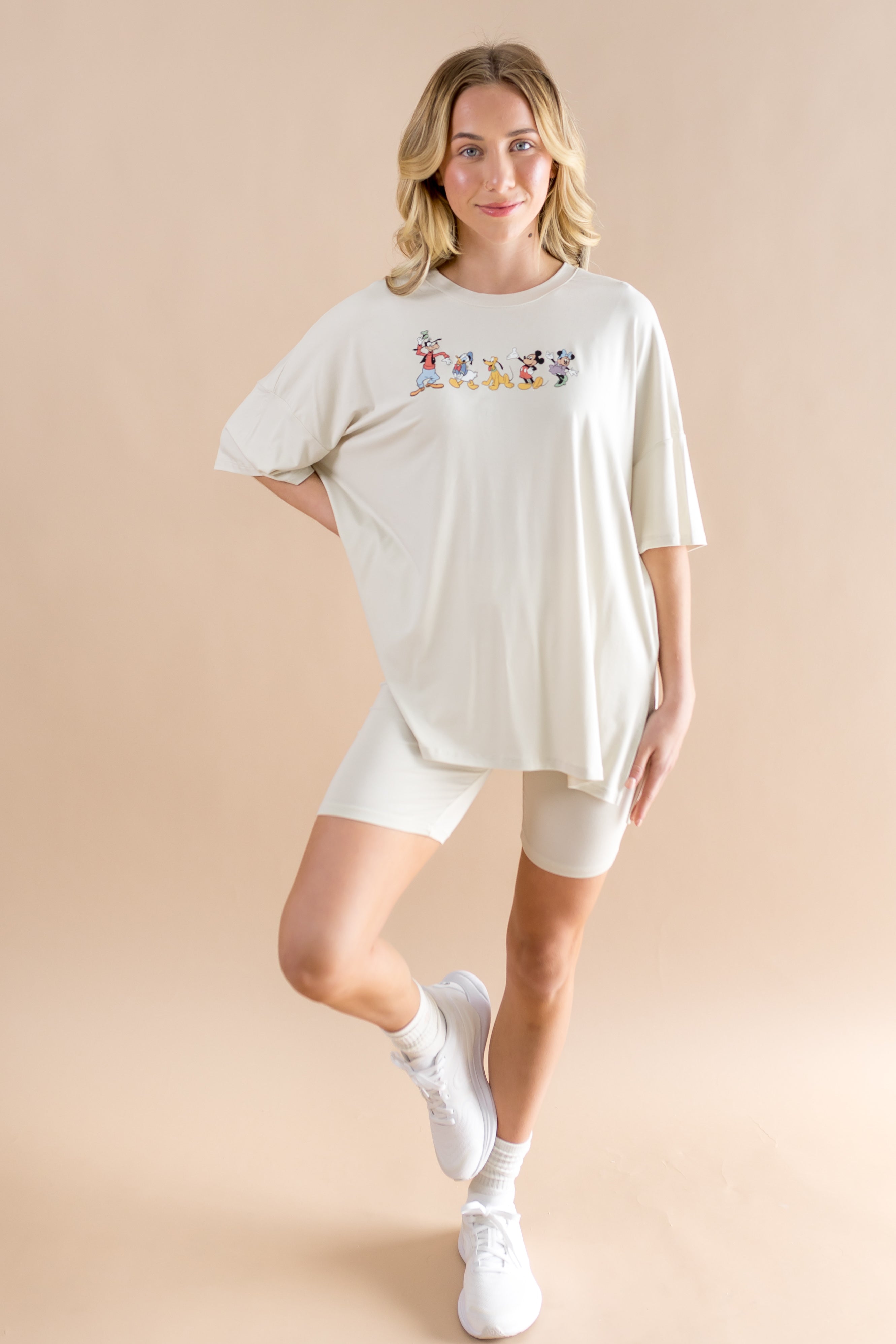 Smiling woman standing with one  leg folded up while modeling the Women’s Biker Short Set in Mickey and Friends on Ecru