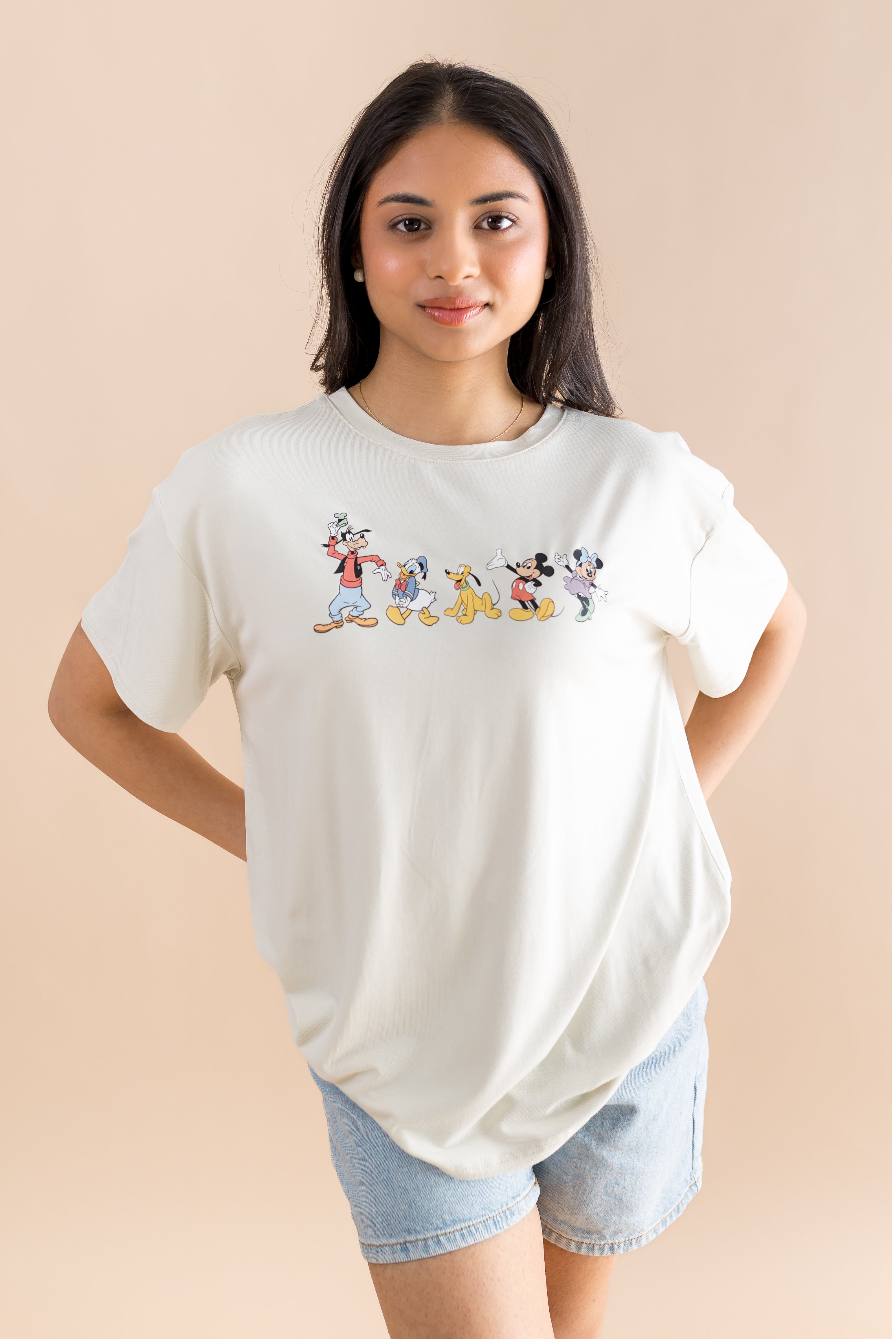 Model standing in front of a beige back ground while wearing the Women's Graphic Tee in Mickey and Friends on Ecru with her hands behind her back paired with jean shorts

::: Model is 5' wearing size XS ///