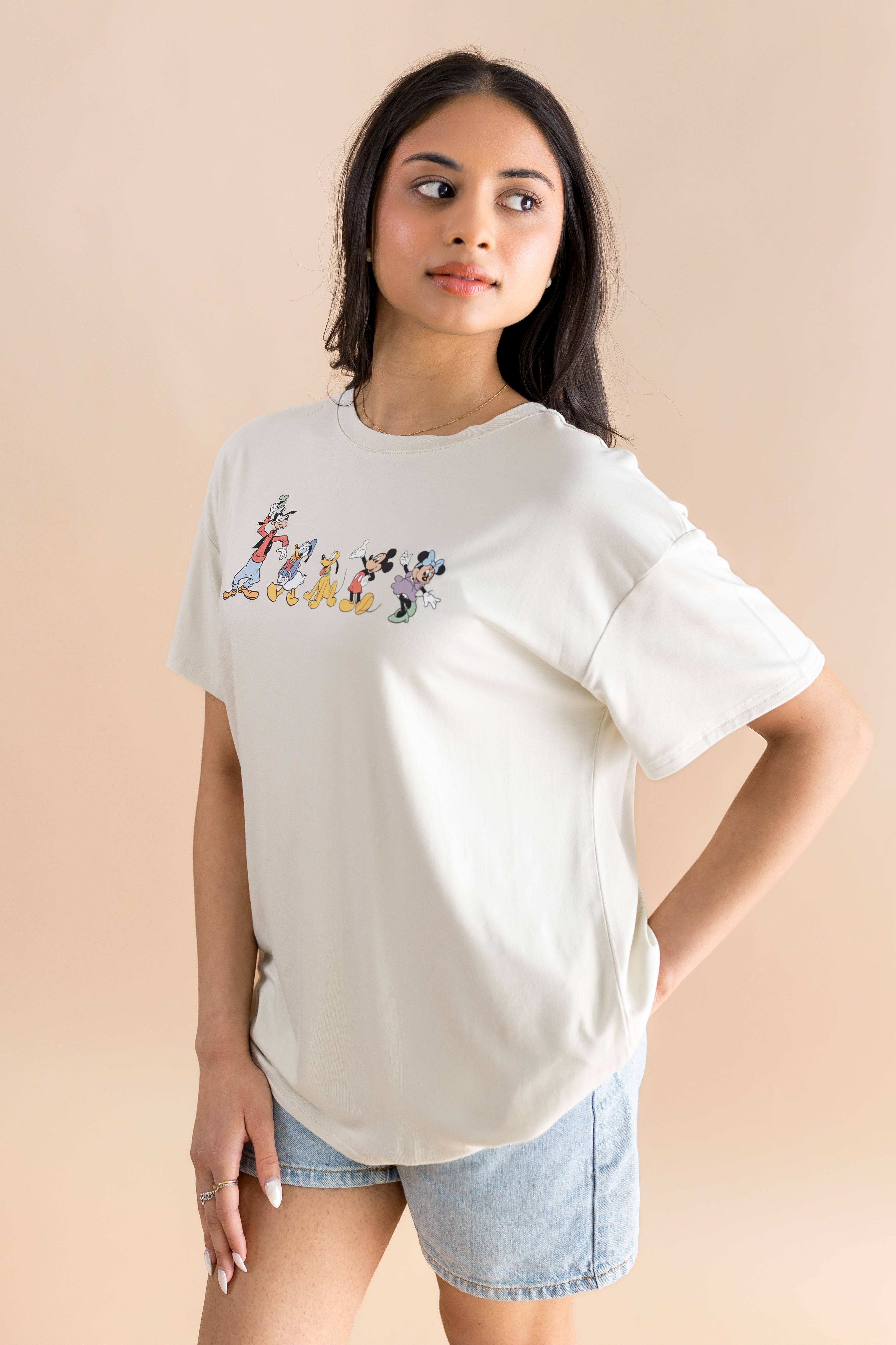 Model standing to the side while wearing the Women's Graphic Tee in Mickey and Friends on Ecru paired with light wash denim shorts